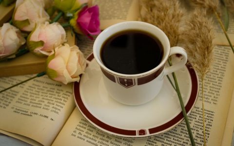 A coffee cup on a saucer rests on an open book surrounded by flowers and reeds, creating a cozy and elegant scene.