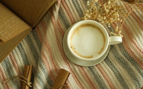 Overhead view of a cappuccino next to books and dried flowers on a woven blanket, creating a cozy scene.
