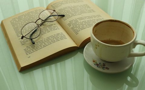 Open book with glasses and used coffee cup on a reflective surface, ideal for literature lovers.