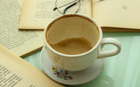 An empty coffee cup surrounded by open books and glasses on a glass table.