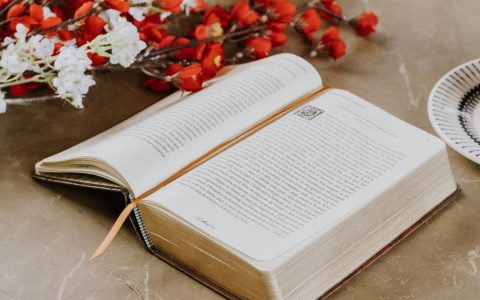 Open book with a bookmark on a table, accompanied by vibrant flowers and a cup.