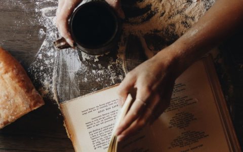 Hands with coffee cup and recipe book on flour-dusted table with bread.