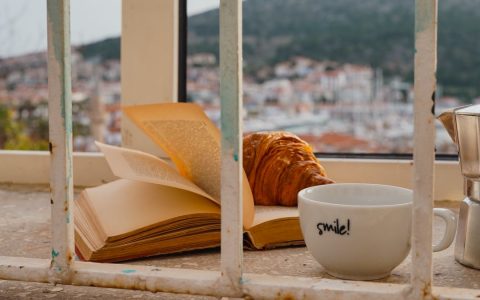 A serene morning scene with coffee, book, and croissant by a window overlooking a scenic town.