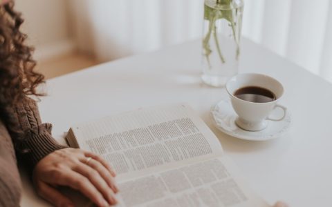 Woman reading a book at a white table with coffee and a vase, creating a cozy moment.