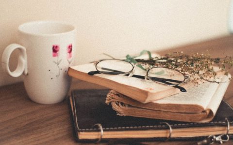 A warm, vintage setup featuring books, glasses, and a floral coffee mug on a wooden table.