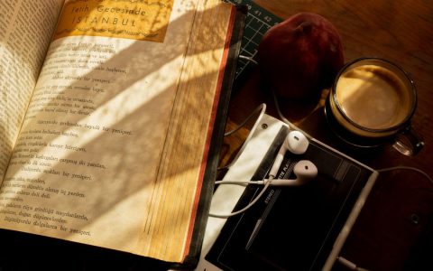 A cozy scene with a vintage book, earphones, coffee, and peach on a wooden desk.