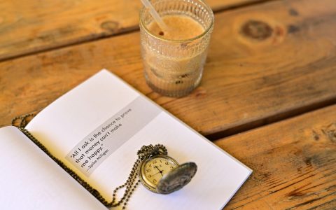Artistic setup of a vintage pocket watch on an open book with a glass of iced coffee on a wooden table.