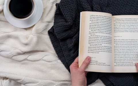 Overhead view of hands holding a book beside a coffee cup on a cozy blanket.