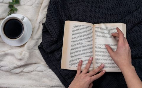 Hands holding an open book with a coffee on a cozy blanket background.