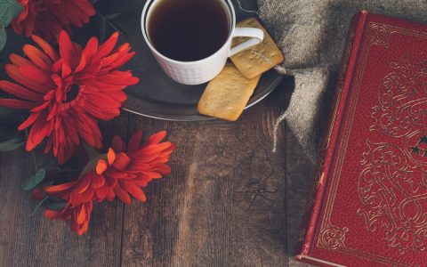 Cozy flat lay with a cup of tea, red flowers, and an ornate book on wooden background.