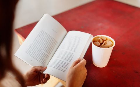 Person reading a book with a cup of iced coffee on a vibrant red table, enjoying a relaxing moment.