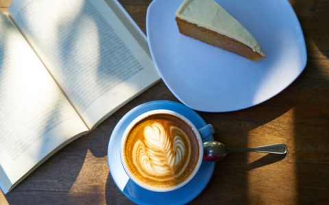 A beautifully crafted latte with latte art, alongside a slice of cake and an open book on a wooden table.