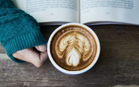 A warm latte with artistic foam held next to an open book on a wooden table, perfect for a cozy moment.