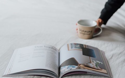 From unrecognizable person sitting on white comfy bed with cup of hot drink and opened magazine
