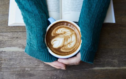 Hands holding a latte with intricate latte art over an open book on a wooden table.