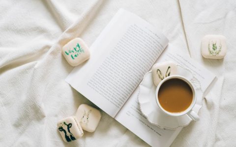 A cozy flat lay with coffee, a book, and Easter-themed macarons on soft white fabric.