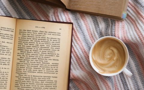 A cup of coffee and open books on a striped blanket evoke a cozy reading atmosphere.
