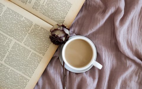 A cup of coffee with an open book and a dry rose on fabric, perfect for a cozy and relaxing ambiance.