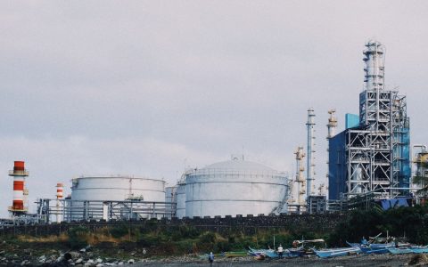 A coastal industrial petrochemical plant with storage tanks and factory structures against a cloudy sky.