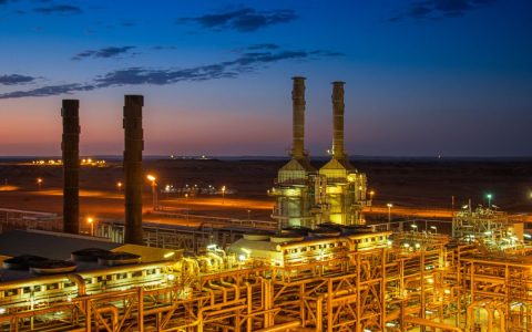 Aerial view of an illuminated factory with chimneys at sunset, showcasing industrial architecture.