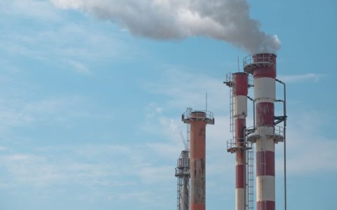 Three industrial smokestacks with white smoke against a clear blue sky, symbolizing air pollution.