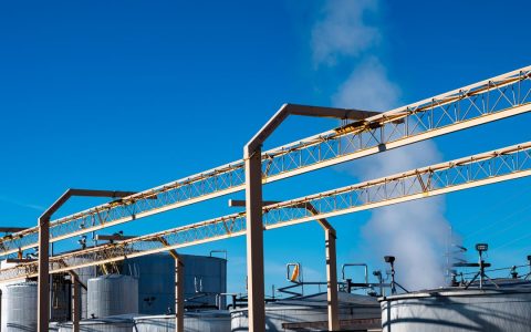 Wide view of an industrial plant with smoke rising from storage tanks against a clear blue sky.