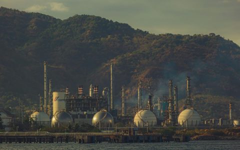 Industrial factory near mountains by a river, showcasing chimneys and infrastructure.