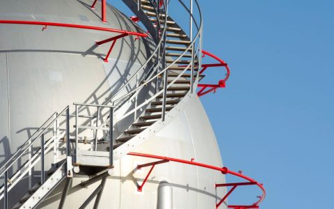 Close-up of a large industrial storage tank with spiral steel stairs against a clear blue sky.
