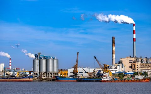 An industrial port scene with smokestacks, silos, and cranes by the water, showcasing urban industry.