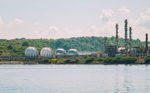 A scenic view of a coastal industrial gas storage facility with spherical tanks and tall chimneys.