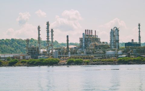 A large oil refinery situated by the water with bright green foliage in the background; clear blue skies and clouds overhead.