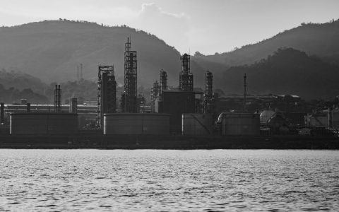 Black and white photo of an industrial refinery with mountains in the background and water in the foreground.