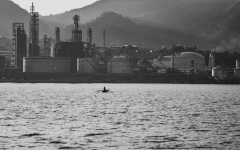 A lone kayaker paddles on a lake with an oil refinery in the backdrop of mountains.