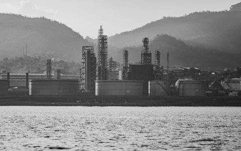 A monochrome shot of an industrial refinery near mountains, highlighting its structures.