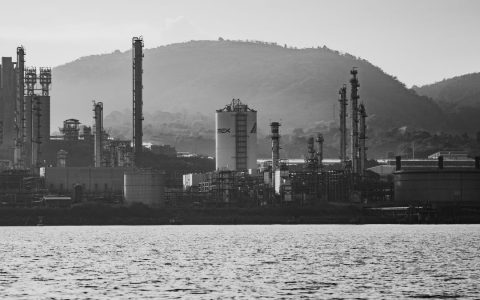 Monochrome view of an industrial complex with hills in the background, captured during the day.