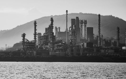 Black and white image of an industrial refinery set against a mountainous backdrop with water in the foreground.