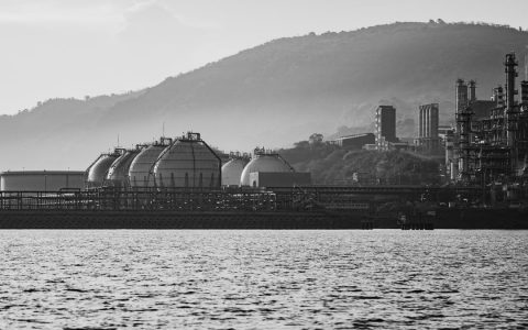 Monochrome image of industrial plant by the sea, framed by hills and water.