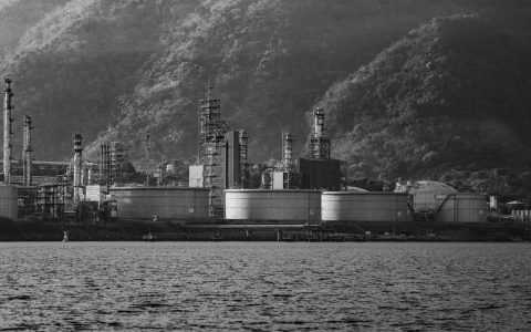 Black and white image of an industrial facility by a mountain range and a body of water.