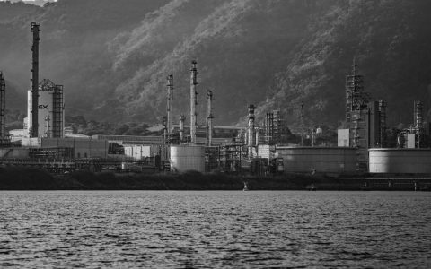 Black and white image of an industrial complex near a water body with mountain backdrop.