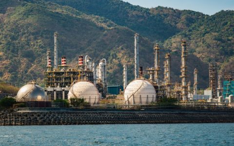 Industrial refinery with spherical tanks by the coastline against a backdrop of mountains.