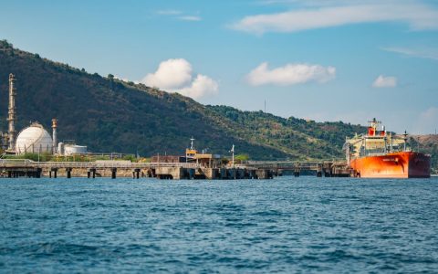 A large oil tanker docked at an industrial port with refinery and mountainous backdrop.