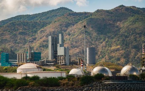 Industrial gas tanks and chimneys against lush hills, showcasing industry and nature.