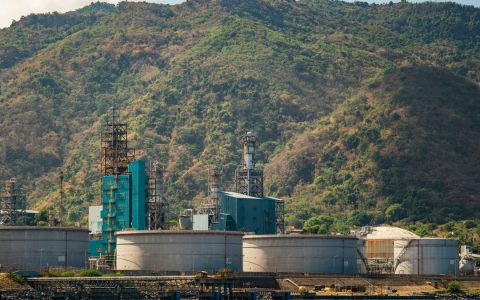 Industrial plant with large tanks and chimneys against a lush mountain backdrop.