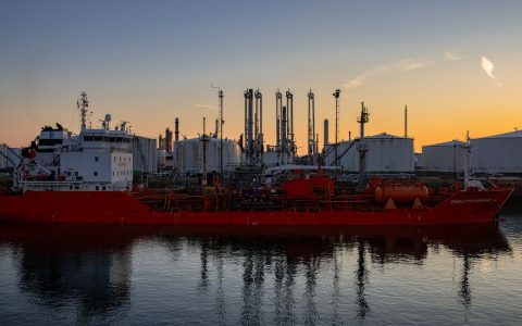 Red cargo ship docked at oil refinery during sunset, showcasing industrial harbor scene.