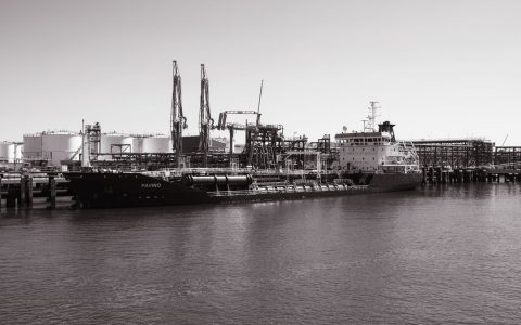 Monochrome photo of a cargo ship docked at an industrial port with cranes in the background.
