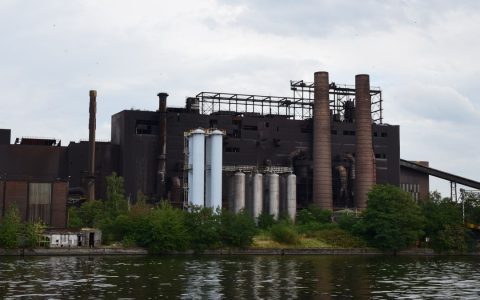 A large industrial factory with chimneys by a river, surrounded by trees on a cloudy day.