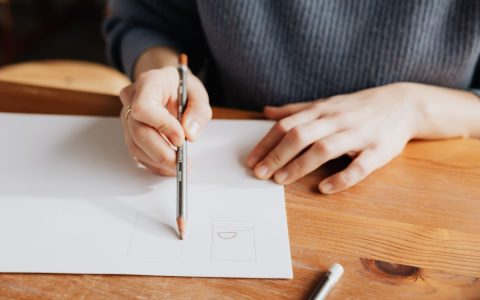 Person sketching on white paper with pencil on a wooden table, showcasing artistic creativity.