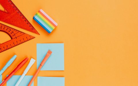 Top view of vibrant stationery essentials including rulers, pens, and sticky notes on an orange background.