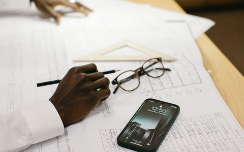 Close-up of an architect drafting on papers with a smartphone and glasses on a desk.