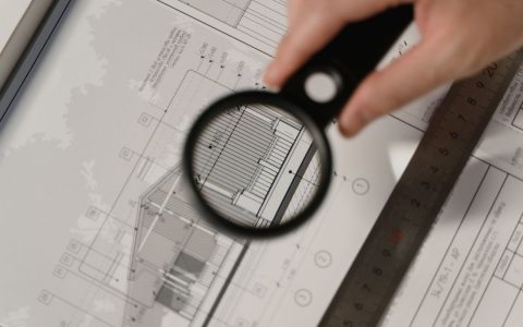 Focused view of architectural plans with magnifying glass, ruler, hands.